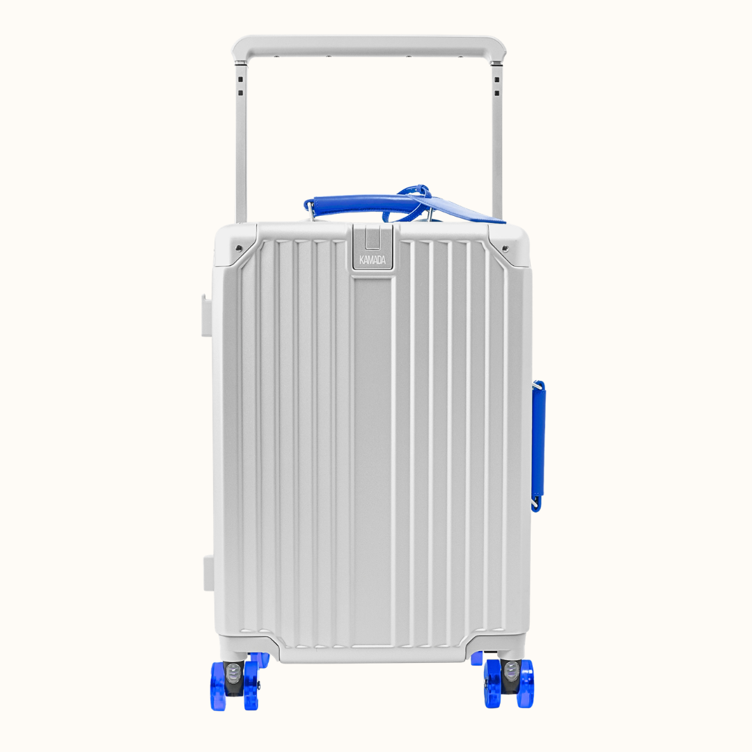 A silver aluminum hard-shell suitcase with vertical ribs, a telescoping handle extended, and bright blue accents on the top handle, side handle, and wheel areas, shown on a plain off-white background.