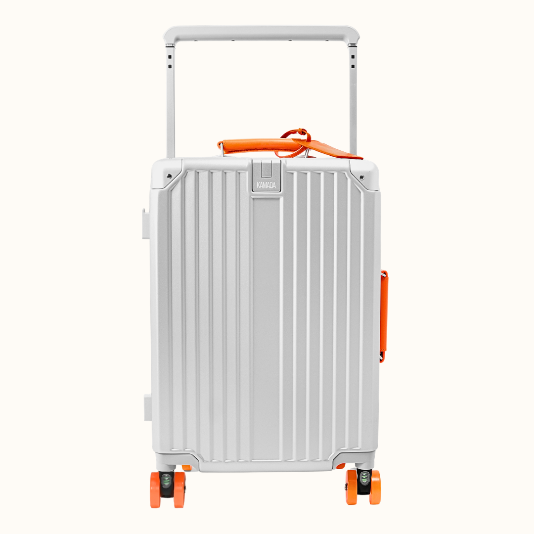 A silver aluminum rolling suitcase with vertical ridges, featuring bright orange accents on the handle, side handle strap, and wheels, shown against a solid off-white background.