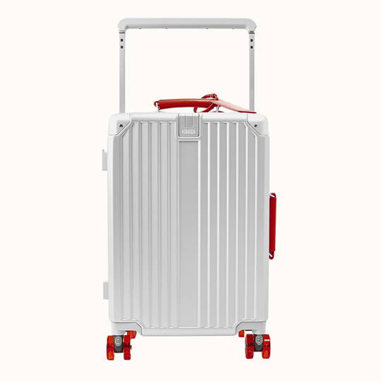 A silver hard-shell carry-on suitcase with vertical ridges, a telescoping handle extended, dual spinner wheels, and red accents including a top handle detail, side handle, and rear reflector, shown on a plain off-white background.