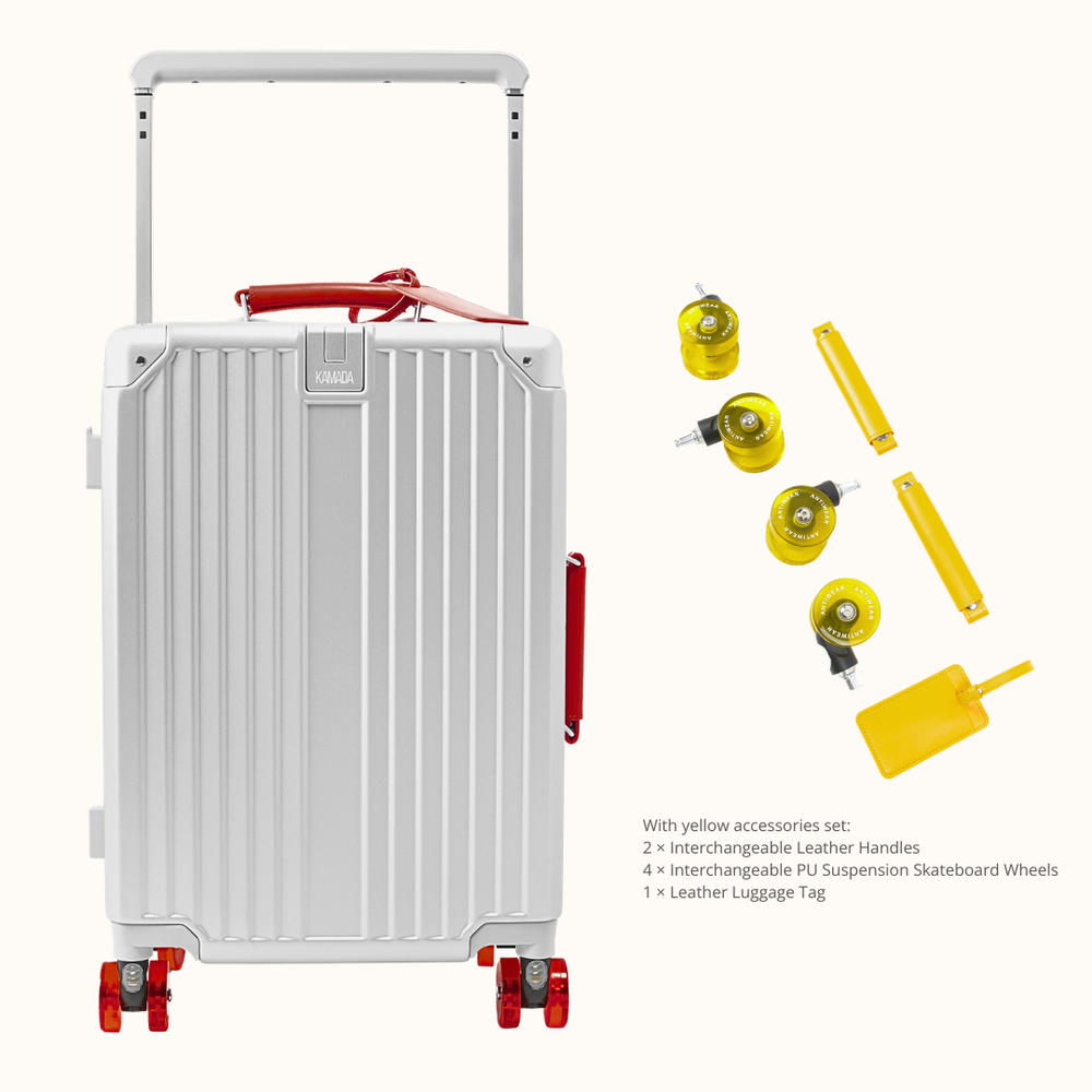 A silver aluminum rolling suitcase with red wheels and a red handle grip, shown alongside yellow accessories including two interchangeable leather handles, four interchangeable PS suspension skateboard wheels, and a leather luggage tag on a light beige background.